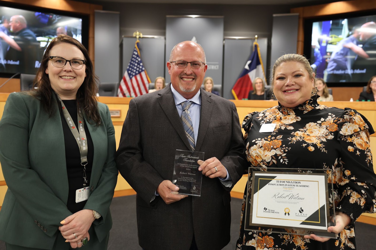 three individuals standing in front of the Plano ISD boardroom, the middle man is holding a certificate and plaque